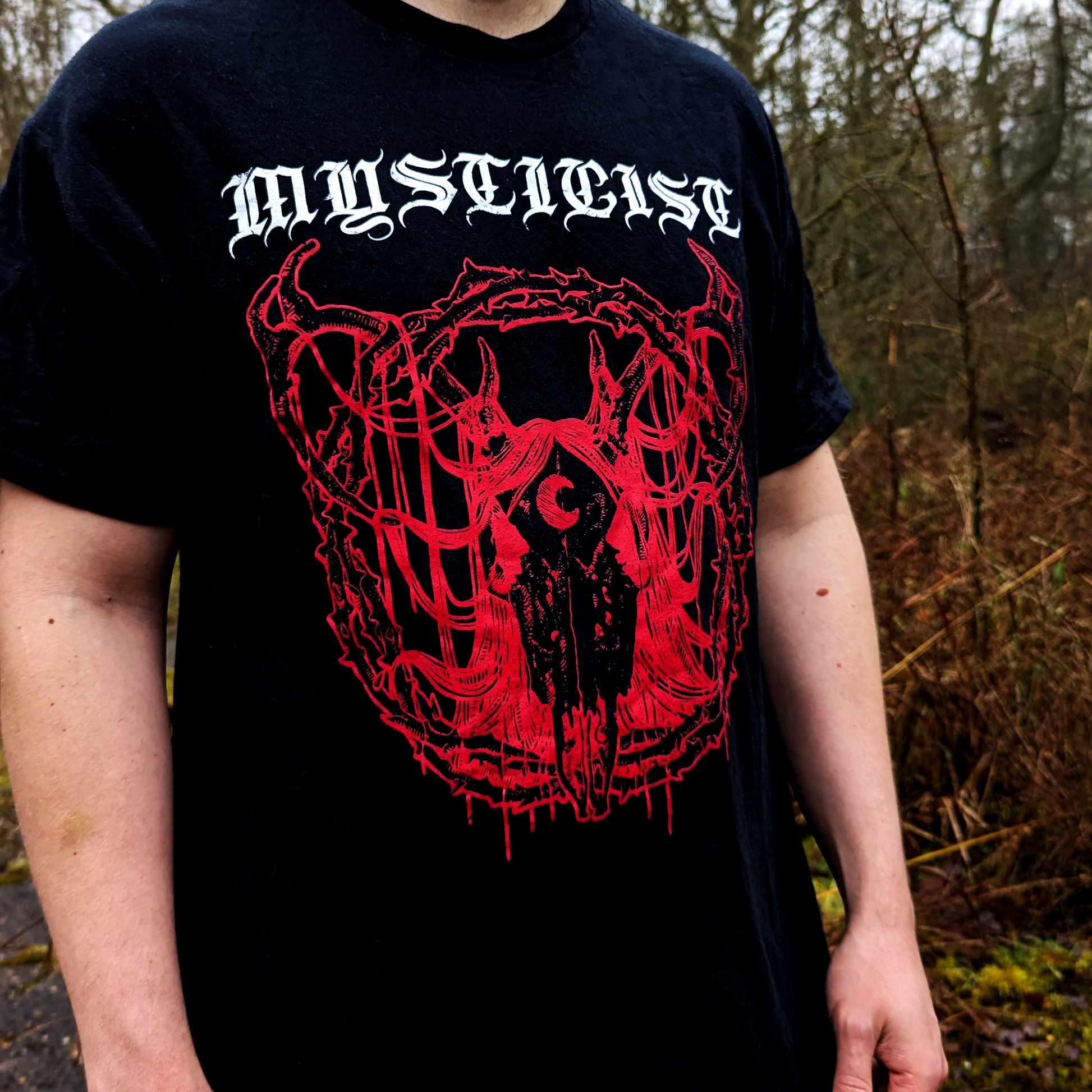 Close up of a black T-shirt worn by a male. Background is a forest. Front of shirt has "Mysticist" band logo and a red design featuring a deer skull with a crescent moon on its forehead. Surrounded by a circle of thorns and covered in long hair draping over its antlers and the circle of thorns.